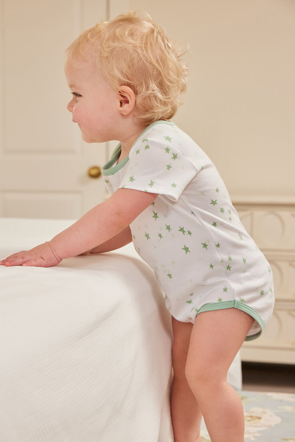 A toddler stands by a bed in a softly lit room, wearing the Baby Bodysuit Bundle in Parisian Green, made from 100% Pima cotton, and looks to the left.