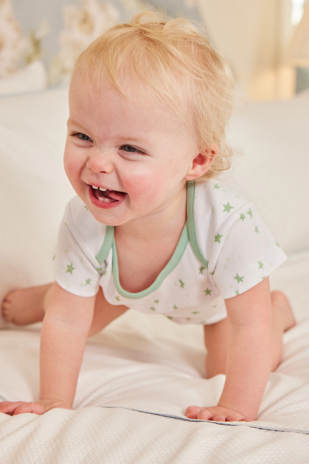 A smiling blonde baby in the Baby Bodysuit Bundle in Parisian Green crawls playfully on a white bed, looking happy and comfortable.