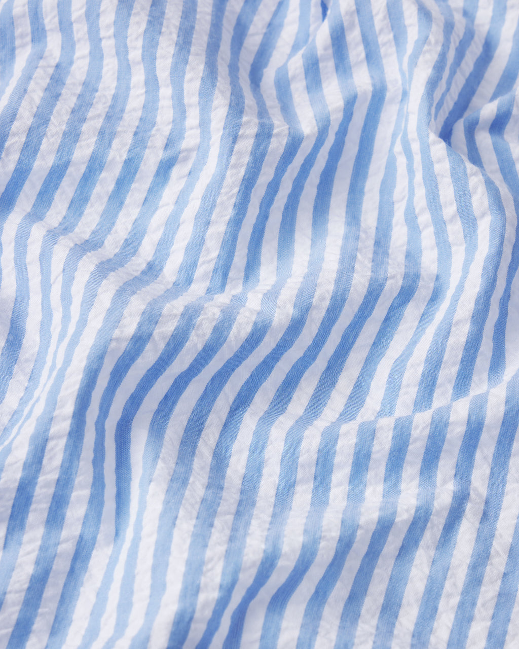 Close-up of light blue and white vertical stripes on crinkled Blue Seersucker fabric from the Georgie Pocket Pants Set, creating gentle waves and shadows across the textured pattern.
