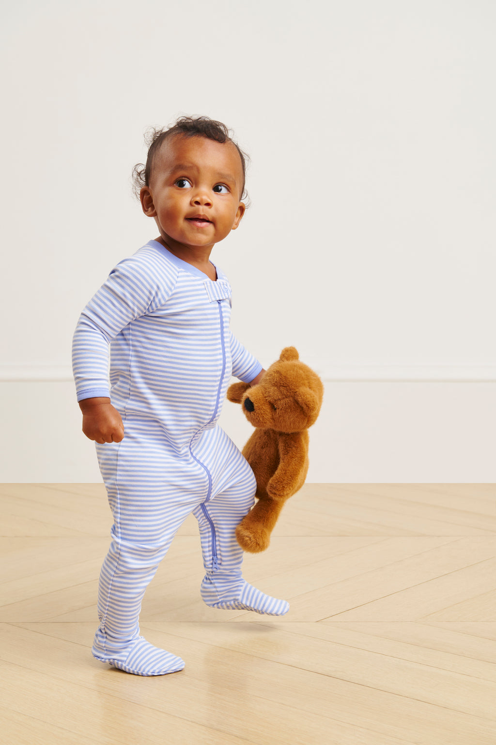 A toddler in the Baby Sleeper in Hydrangea, featuring blue and white stripes and a dual zipper, stands on a wooden floor holding a brown teddy bear and looking up with curiosity.