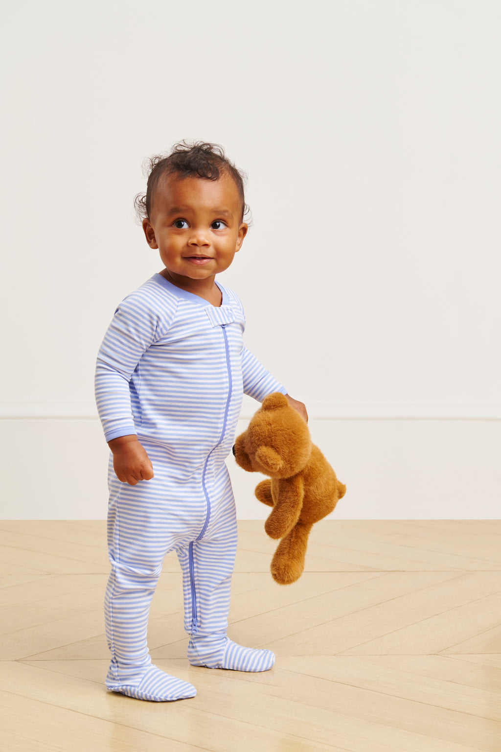 A toddler wearing the Baby Sleeper in Hydrangea stands on a wooden floor, holding a brown teddy bear and smiling slightly against a plain light background.