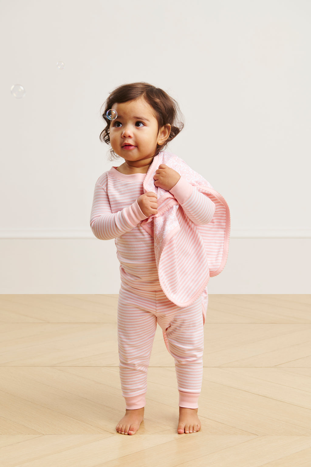 A toddler wearing the Baby Long-Long Set in English Rose Stripe holds a matching blanket, standing on a light wood floor. A soap bubble drifts near their face against an off-white wall.
