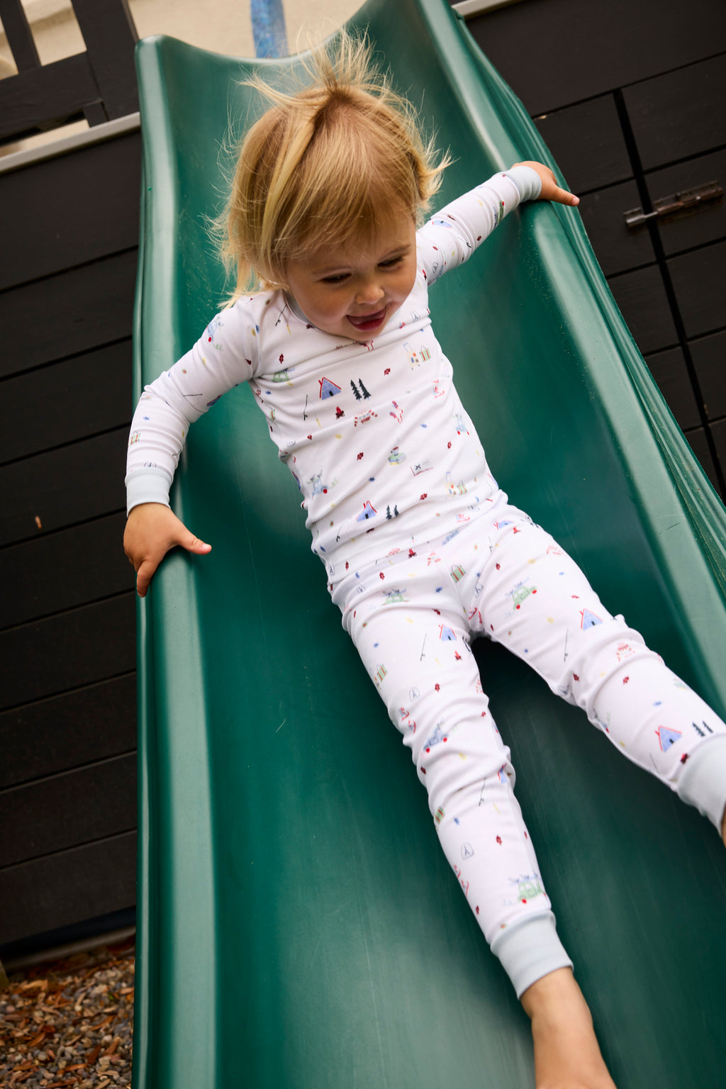A young child in the Kids Long-Long Set in Alpine Adventure, white pajamas with colorful patterns, slides down a green playground slide smiling and arms outstretched, ready for an adventure.
