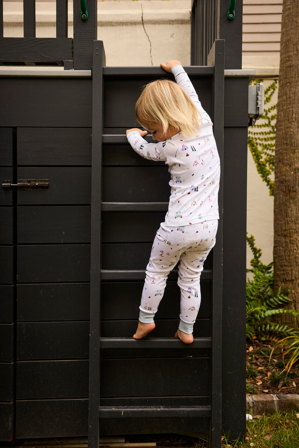 A young child, facing away from the camera, climbs a black wooden ladder outdoors in the Kids Long-Long Set in Alpine Adventure—cozy Pima cotton pajamas. Greenery can be seen nearby.