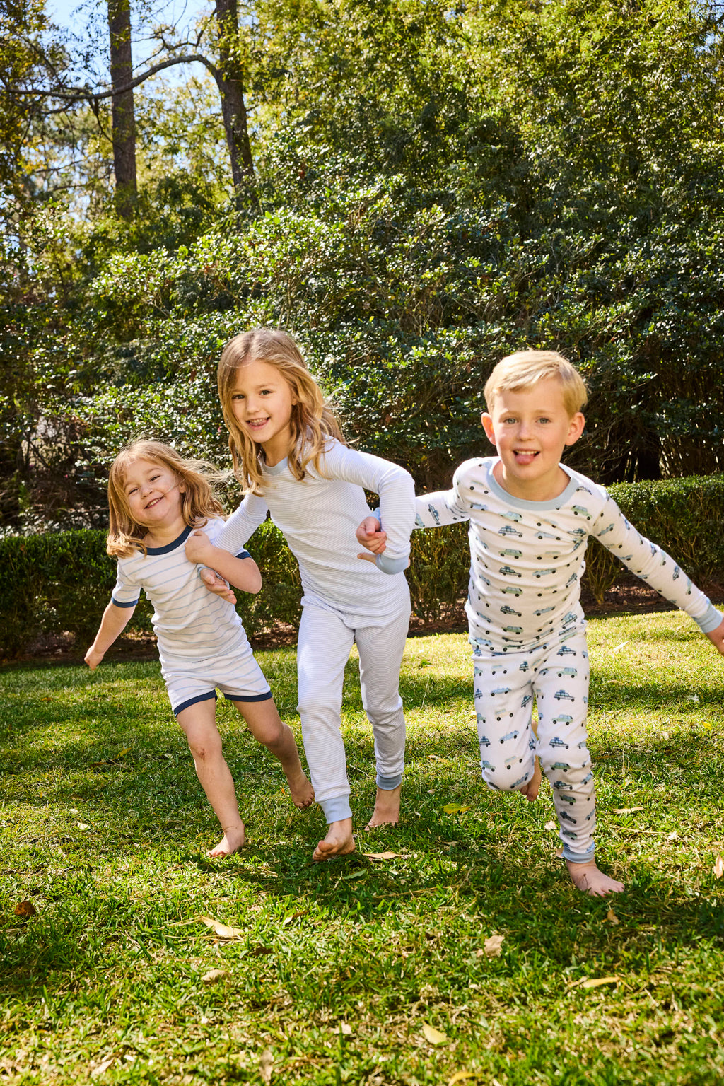 Three kids wearing Kids Long-Long Set in Off Road Cruisers laugh and play on green grass in a sunny backyard, surrounded by trees and bushes.