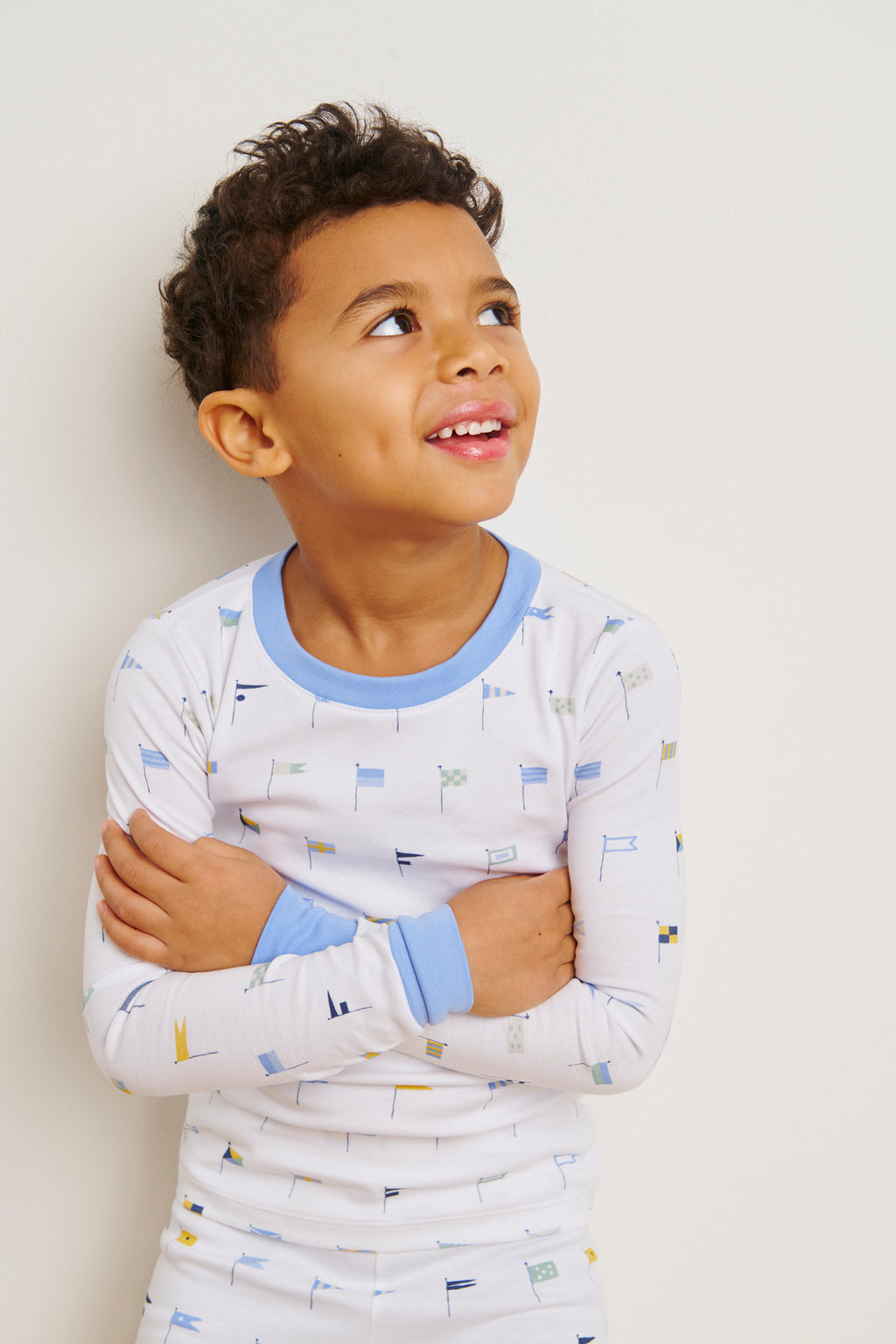 A young boy smiles and looks up with arms crossed, wearing the Kids Long-Long Set in Baltic Blue Nautical Flags—soft Pima cotton pajamas with a light blue collar and cuffs—against a plain light background.