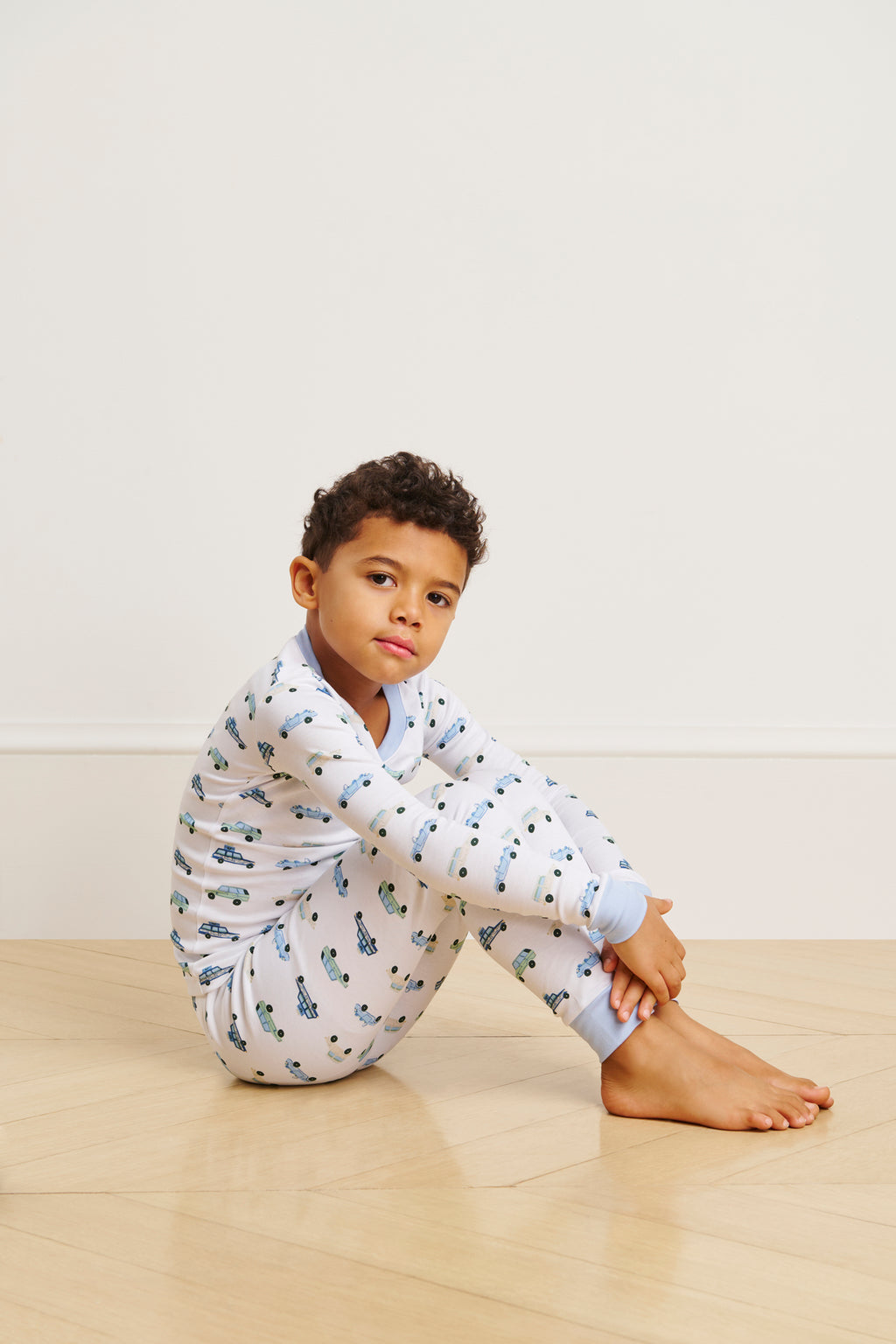 A young child sits on a wooden floor wearing the Kids Long-Long Set in Off Road Cruisers, featuring soft Pima cotton with blue cuffs and a car pattern, gazing at the camera against a plain light background.