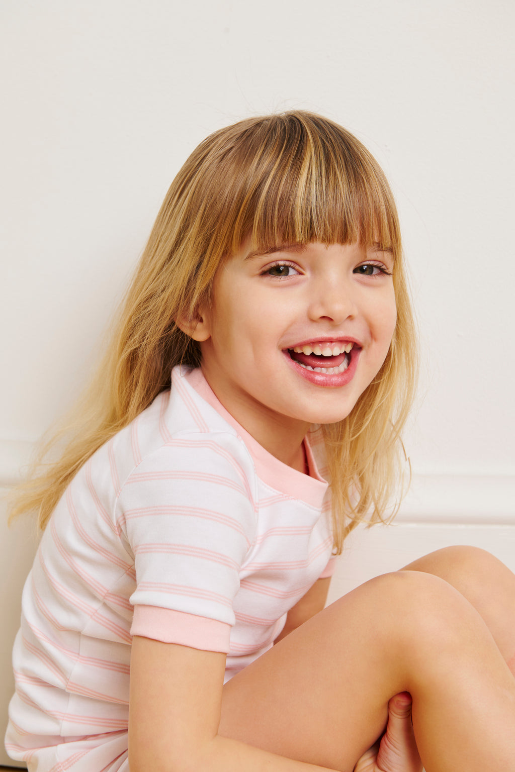 A young child wearing the Kids Shorts Set in English Rose Triple Stripe sits smiling and looking at the camera against a light-colored background.