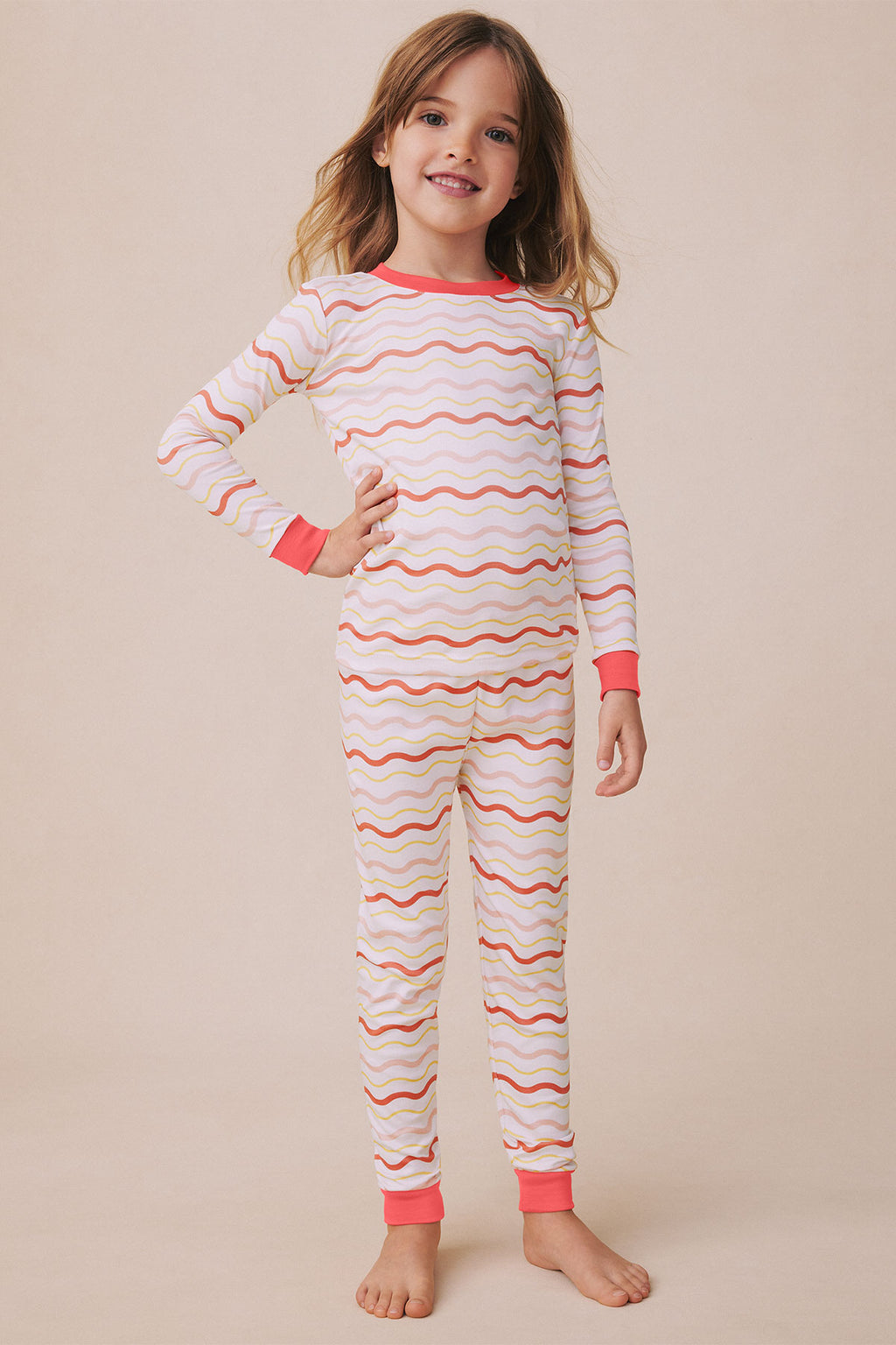 Child wearing long-sleeve pajama set with wavy striped pattern in coral, yellow, and pink with matching pants and cuffed wris