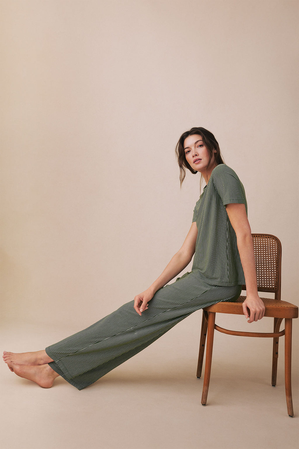 Woman wearing rosemary green short-sleeve top and long pants with vertical micro stripe pattern, seated on wooden chair.