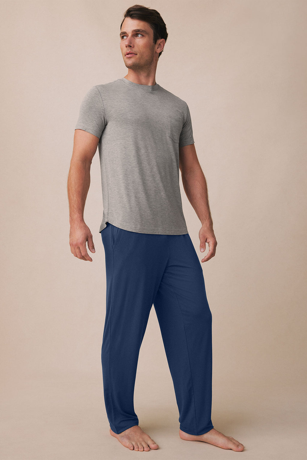 Man wearing gray short-sleeve t-shirt and navy pants, standing barefoot against neutral background.