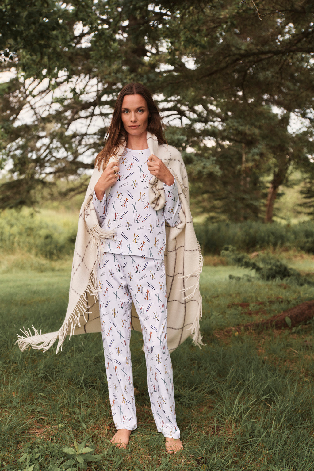 A woman stands on grass with trees behind her, wrapped in a blanket and wearing the Pima Crew Long-Long Set in Vintage Skis—light-colored, matching Pima Cotton pajamas with a playful pattern.