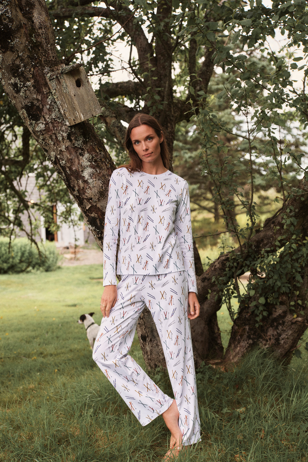 A woman wears the Pima Crew Long-Long Set in Vintage Skis, made from soft Pima Cotton, as she stands under a tree with a birdhouse in a grassy garden while a small dog plays nearby.