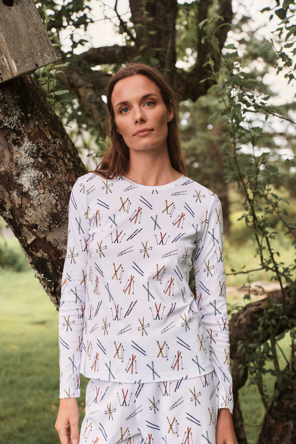 A woman stands outdoors near a tree, wearing the Pima Crew Long-Long Set in Vintage Skis—white long sleeve pajamas with colorful crossed vintage skis—her cozy sleepwear highlighted by the lush green background.