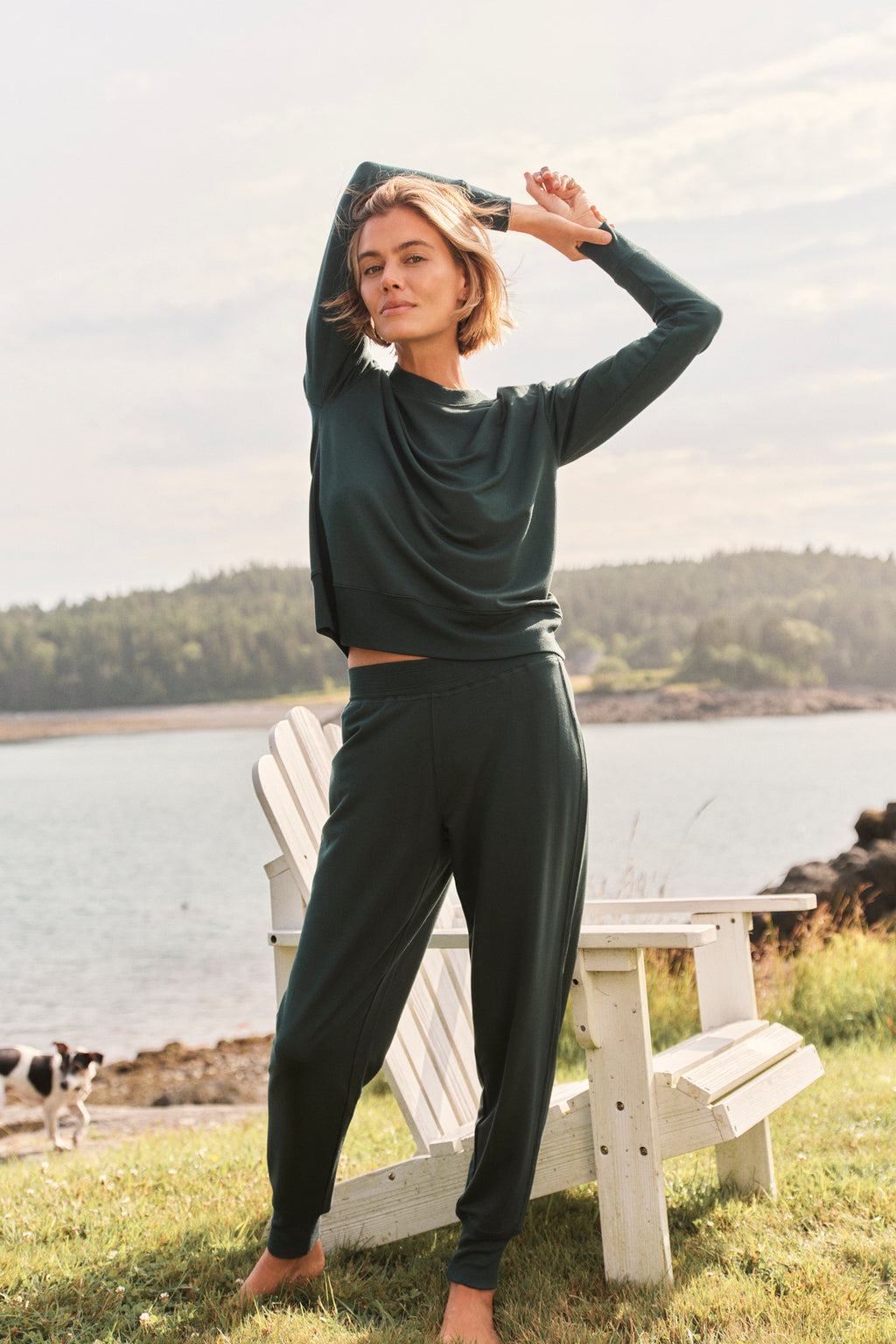 A woman wearing the Relax Jogger Set in Conifer stands on grass near white Adirondack chairs by a lake, with trees and hills behind her. She raises one arm, appearing relaxed, while a small dog is visible in the distance.
