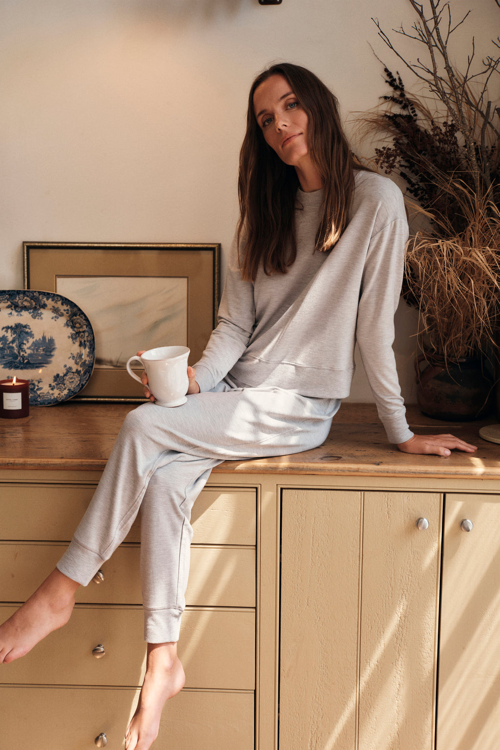 A woman wearing the Relax Jogger Set in Heather Gray sits on a wooden cabinet, holding a white mug. Dried plants, a framed painting, a decorative plate, and a candle decorate the background as warm sunlight streams in.