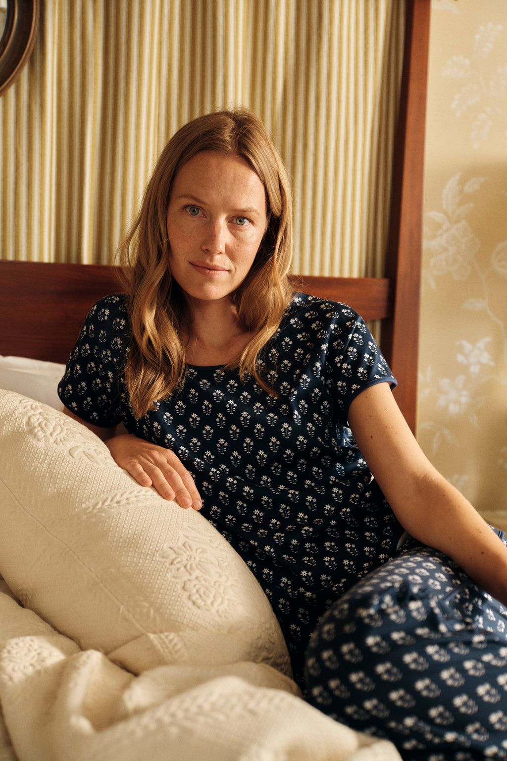 A woman wearing the Pima Short-Long Set in English Navy Posy sits on a bed, leaning against a white textured pillow. The room has striped wallpaper and a wooden headboard.