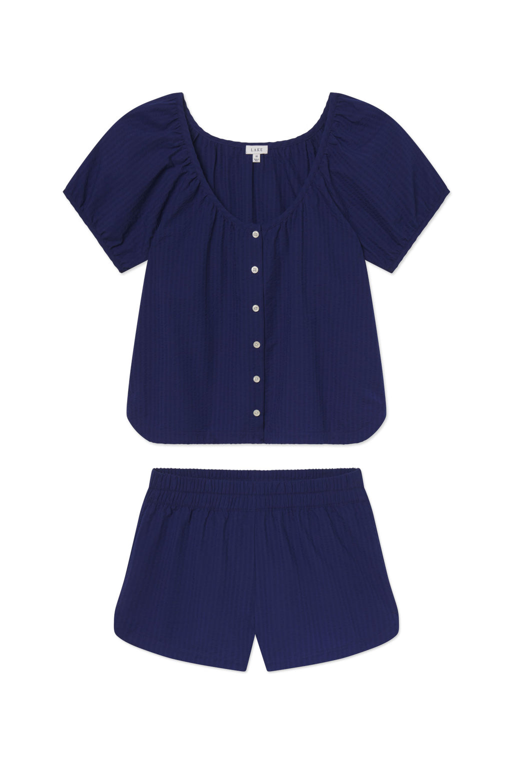 The Charlie Shorts Set in Blue Depths Seersucker is a two-piece navy pajama set with a short-sleeve button-up top, matching shorts, a soft scoop neckline, elastic waistband, and lightweight Pima cotton seersucker fabric.