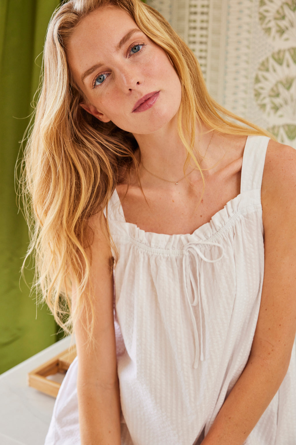 A woman models the Alice Nightgown in White Seersucker, featuring ruffled sleeves, as she stands indoors before a green curtain and a patterned white wall.