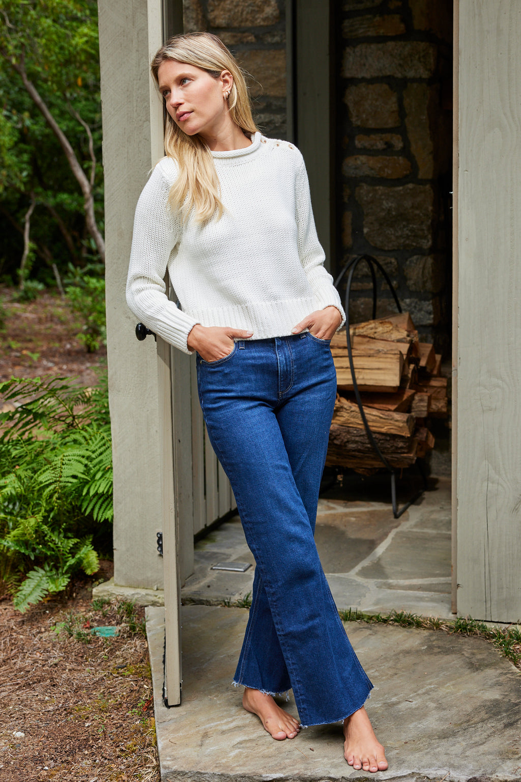 A woman leans against a doorway outside, wearing a chunky knit Jane Cotton Sweater in Ivory and blue flared jeans, with stacked firewood and greenery in the background.