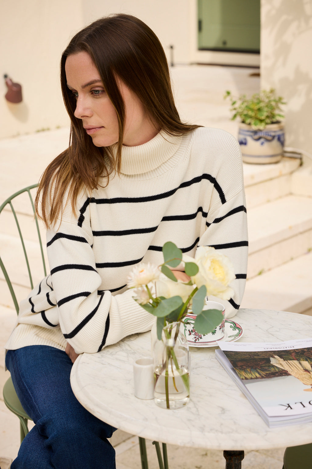 Wearing the Madeline Sweater in Ivory Black Stripe, a woman sits at an outdoor table with a vase of flowers, a teacup, and a magazine, gazing to the side as sunlight brightens the scene.