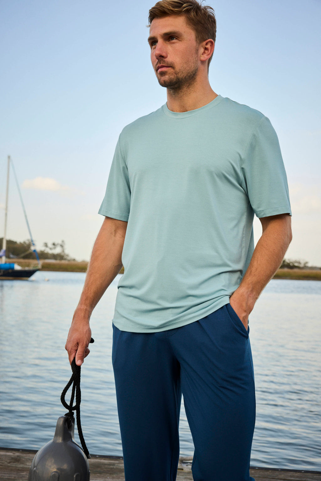 A man stands by calm water holding a boat fender. He wears the Men