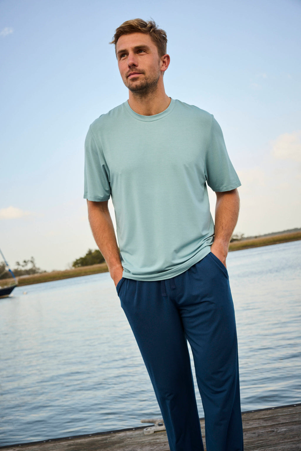 A man stands on a dock by the water, hands in his pockets, wearing the Men