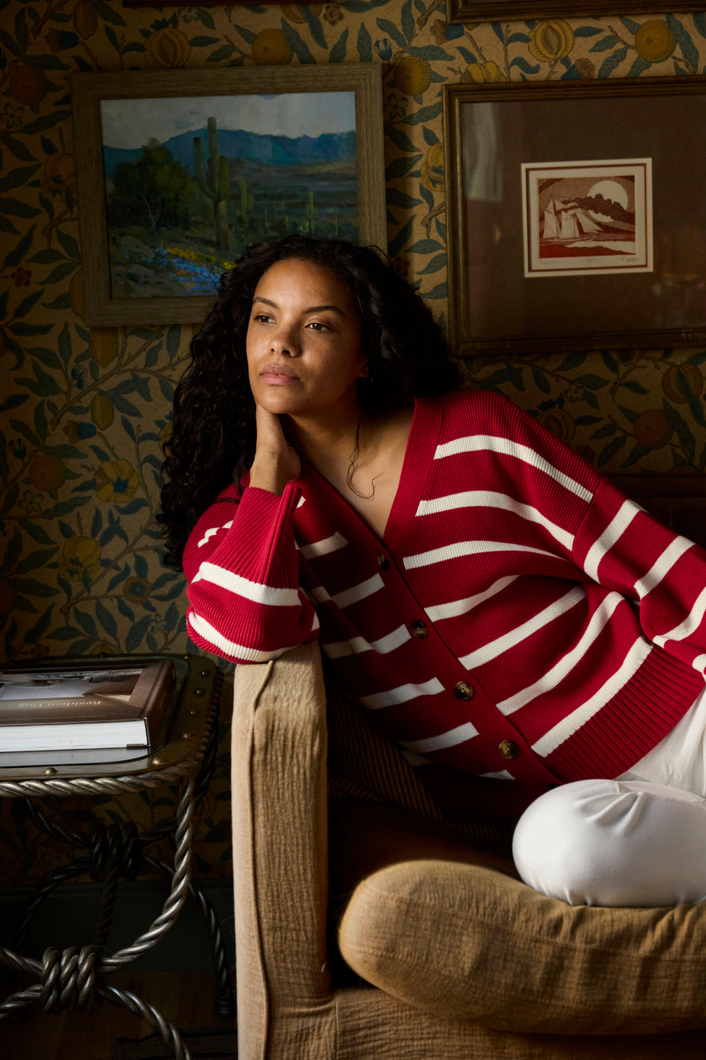 A woman in the Nora Cardigan in Ivory Crimson Inverse Stripe and white pants leans thoughtfully on an armchair in a cozy room with patterned wallpaper and framed art.