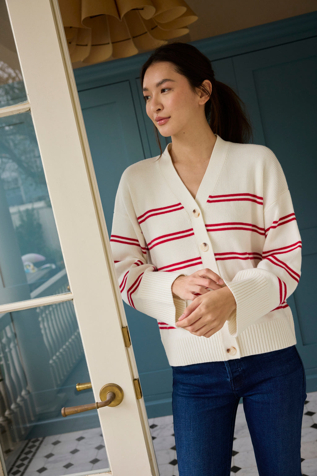 A woman stands indoors by a glass door, wearing the Nora Cotton Cardigan in Ivory Red Stripe with blue jeans. The room has blue walls and a patterned tile floor as she looks to the side.