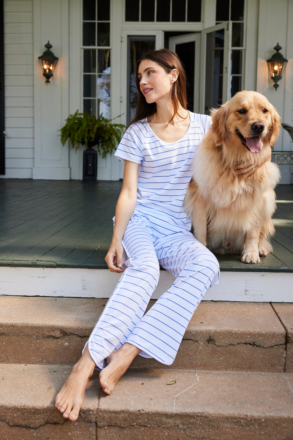 A woman in the Pima Short-Long Set in Cobalt lounges on porch steps beside a golden retriever, both at ease. Behind them stands a white house with green doors, lush plants, and classic wall lanterns.