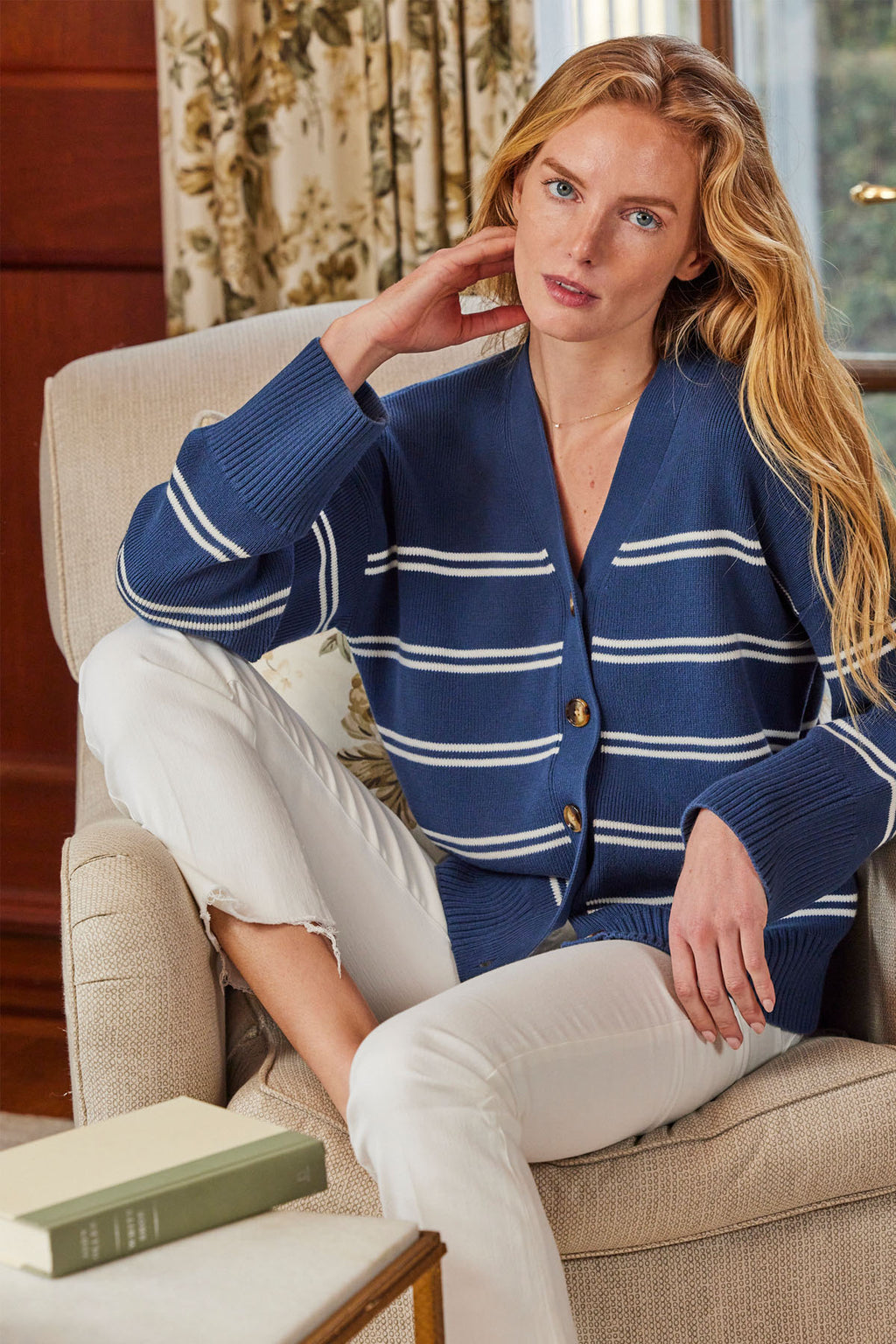 A woman sits on a beige armchair, wearing the Nora Cotton Cardigan in Navy Ivory Stripe with white pants. She rests one hand on her neck and looks at the camera. A green book lies on a wooden table nearby.