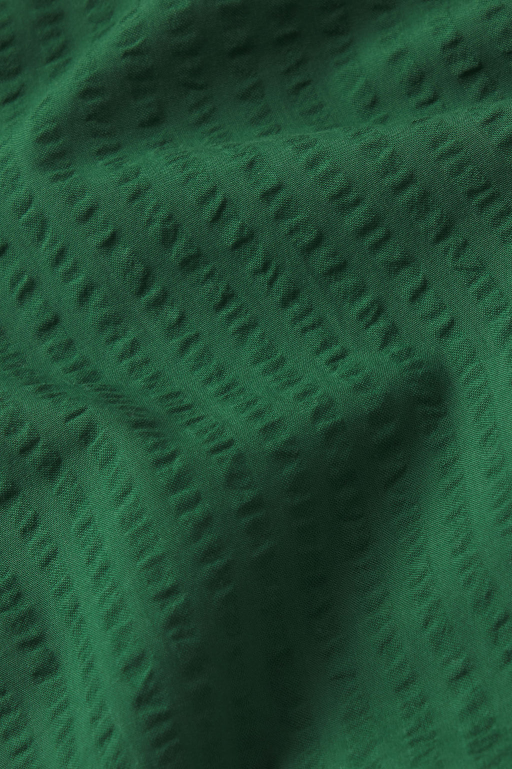 Close-up of the Charlie Nightgown in Eden Seersucker, showing deep green fabric with a subtle, raised dotted pattern running diagonally, creating gentle folds and soft shadows on the surface.