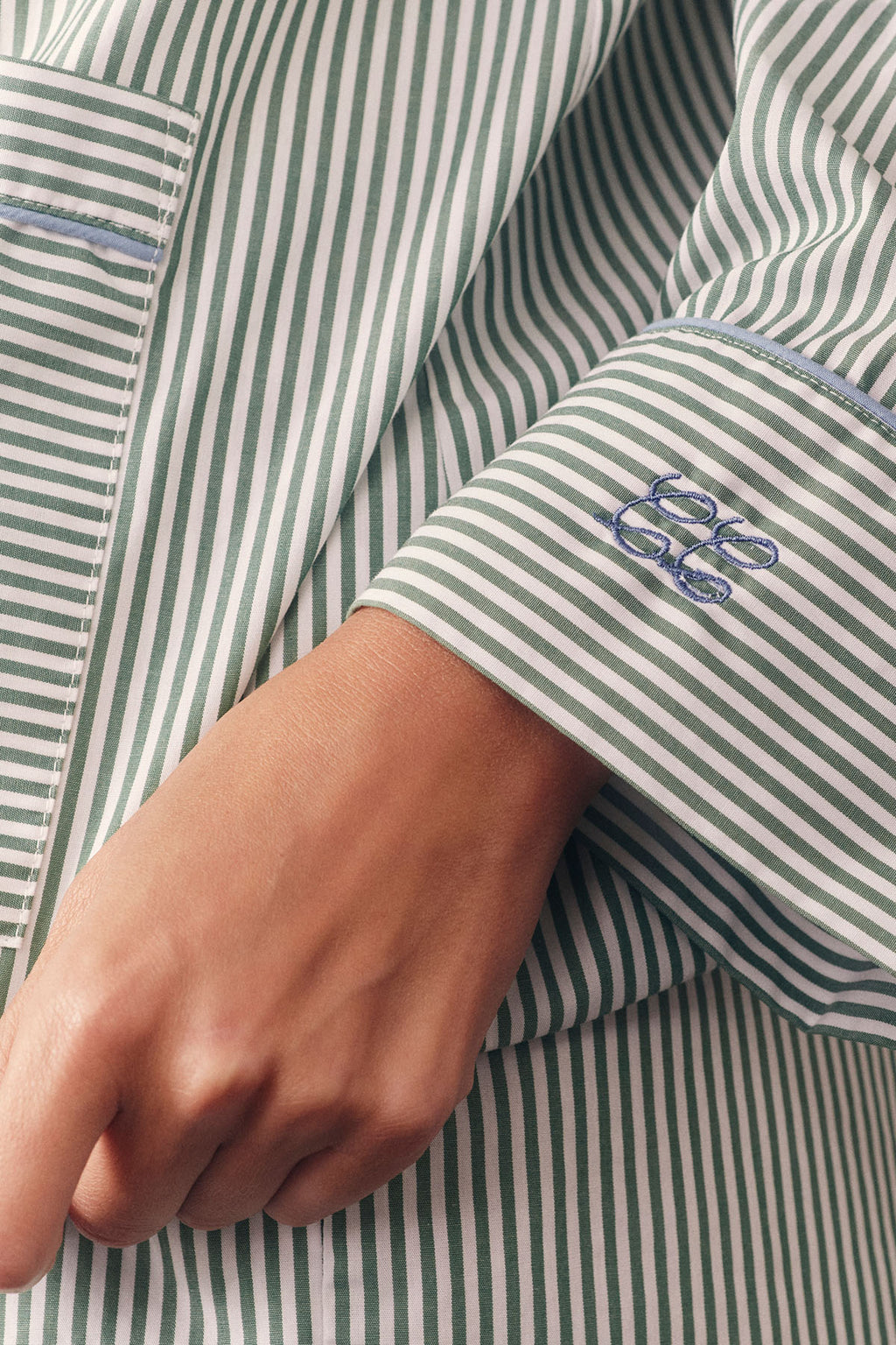 A close-up of a person wearing the Poplin Piped Long-Short Set in Palm Riviera Stripe, featuring green and white stripes, blue “bb” embroidered on the cuff, and a chest pocket. Their hand is resting by their side.