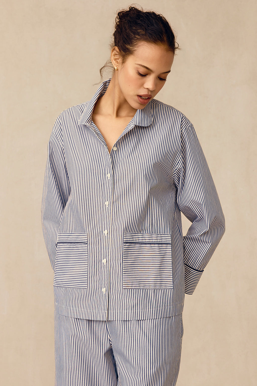 A person models the Poplin Piped Long-Long Set in English Navy Riviera Stripe, featuring blue and white stripes, a collared top with large front pockets, and stands against a beige background looking down with hands behind their back.