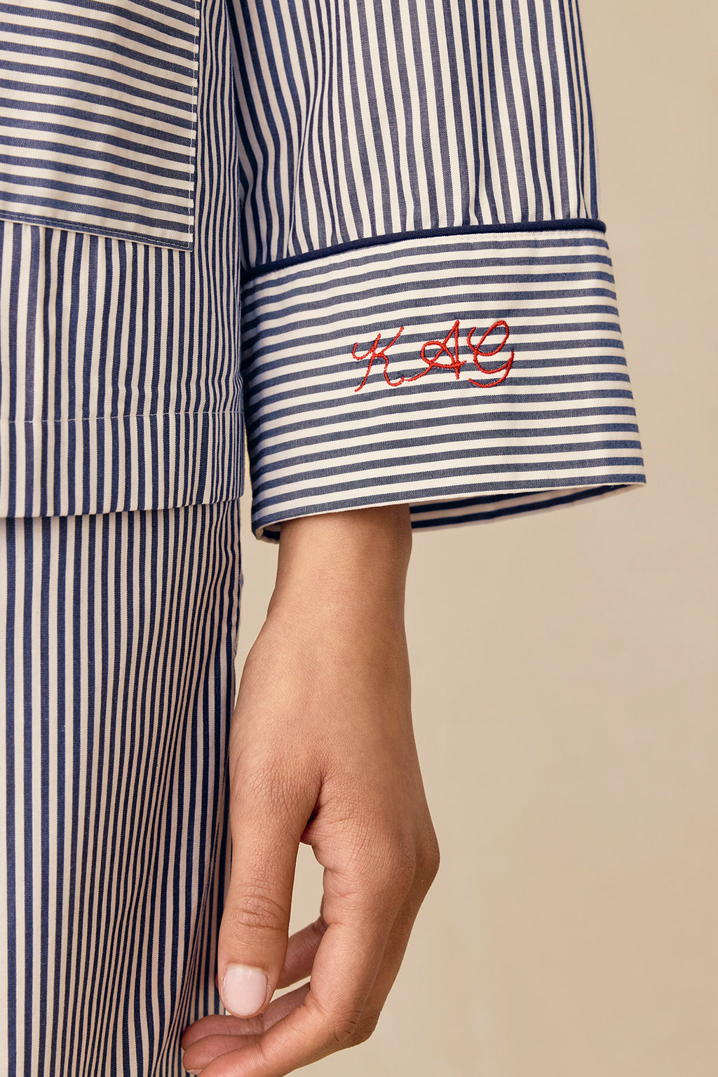 A close-up of a person wearing the Poplin Piped Long-Long Set in English Navy Riviera Stripe shows their arm with a cotton sleepwear shirt cuff featuring red embroidered initials "KAG" and their hand relaxed at their side.