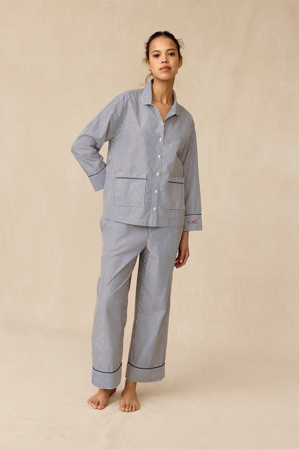 A woman, hair tied back and looking relaxed, stands on a neutral background wearing the Poplin Piped Long-Long Set in English Navy Riviera Stripe—loose-fitting pajamas featuring blue and white stripes, large front pockets, and rolled-up cuffs.