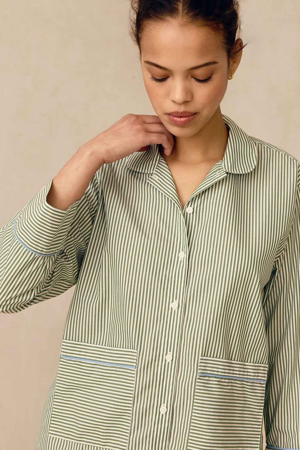 A woman wearing the Poplin Piped Long-Long Set in Palm Riviera Stripe, a green and white button-up pajama with large front pockets, looks downward and gently touches her collar against a plain neutral background.