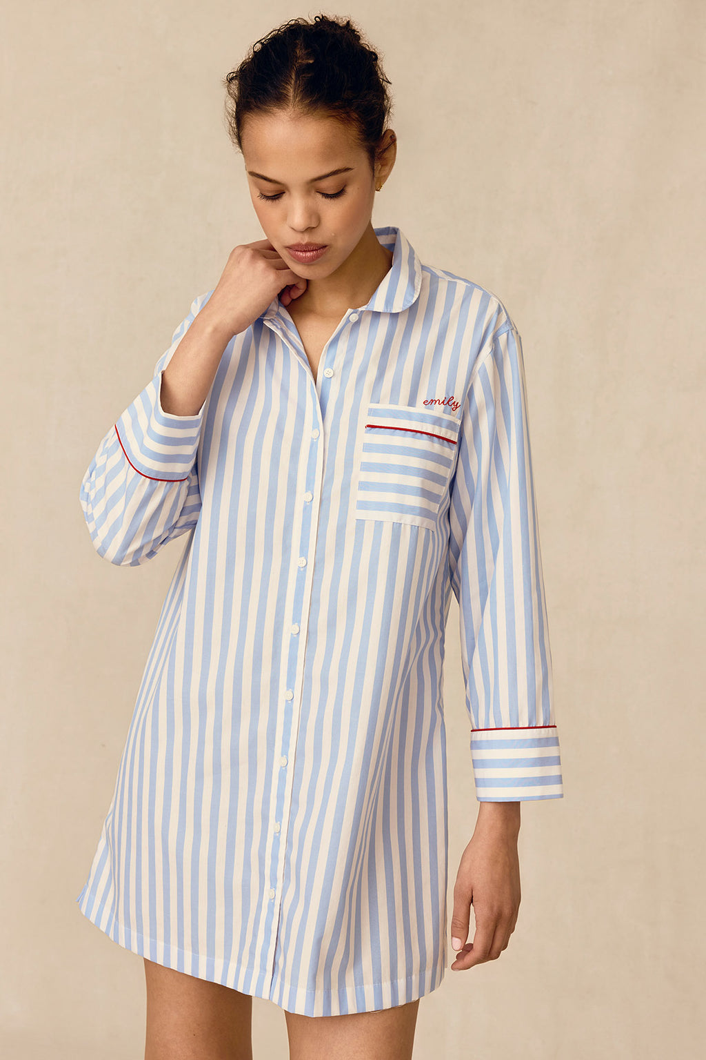A woman wearing the Poplin Piped Nightgown in Baltic Blue Beam Stripe, featuring red accents, stands against a beige background, looking down with one hand touching her neck.