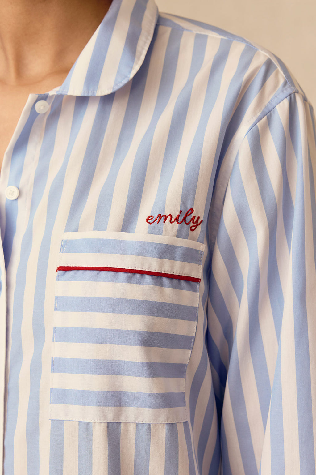 A close-up of a person wearing the Poplin Piped Nightgown in Baltic Blue Beam Stripe, with "emily" embroidered in red above the chest pocket, showcasing its classic style.
