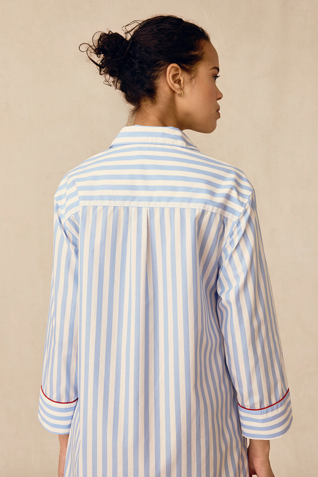 A woman stands with her back to the camera in a Poplin Piped Nightgown in Baltic Blue Beam Stripe, featuring red-trimmed cuffs, against a neutral background.